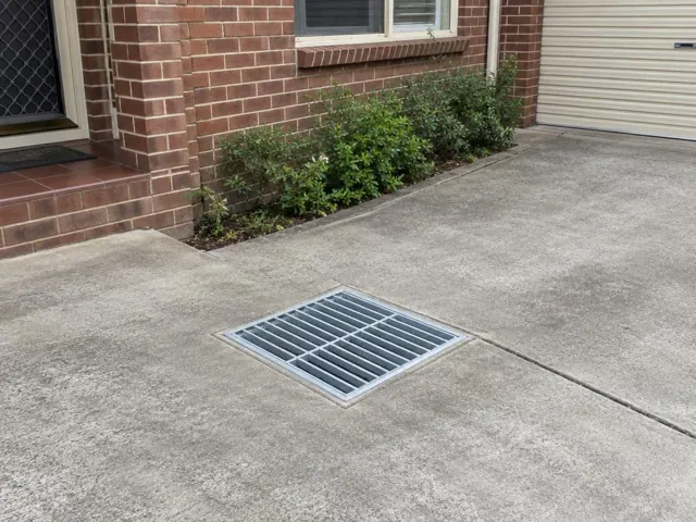 Dry outdoor drain grate set in concrete driveway beside brick house and shrubs in Granville